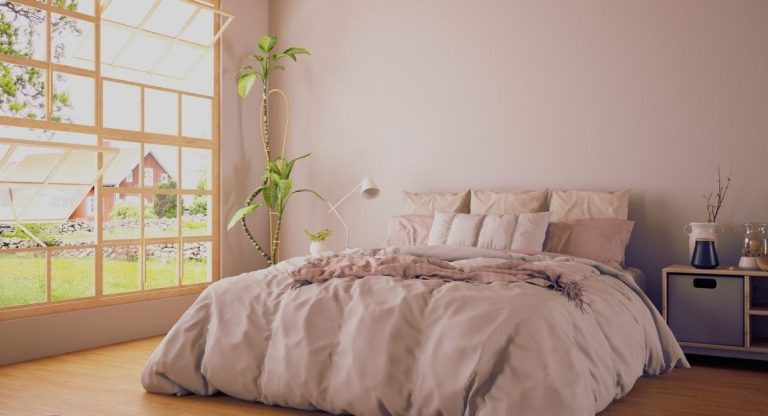 Lit garni de coussins et linge rose poudré, lampe de chevet et plante verte près d'une grande fenêtre